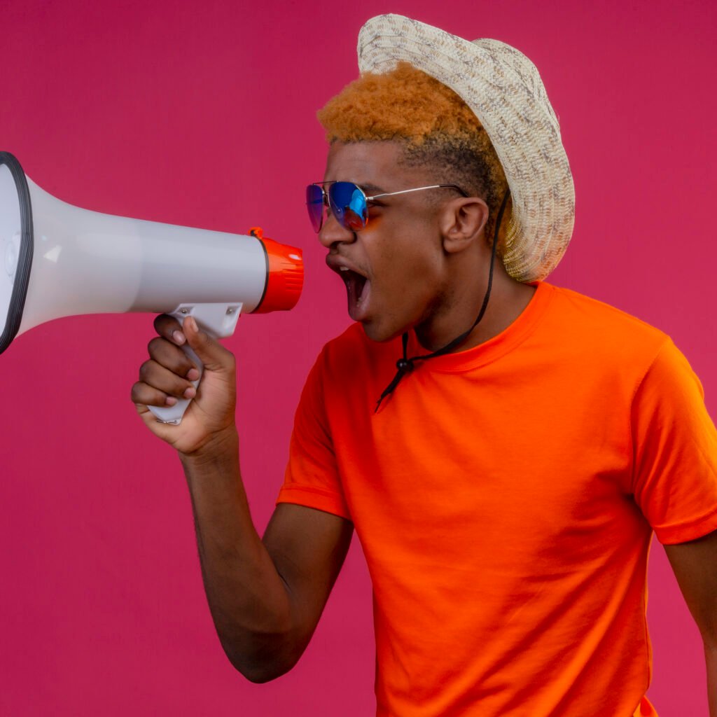 young african american handsome boy in summer hat wearing orange t-shirt shouting to megaphone with angry expression standing over pink background
